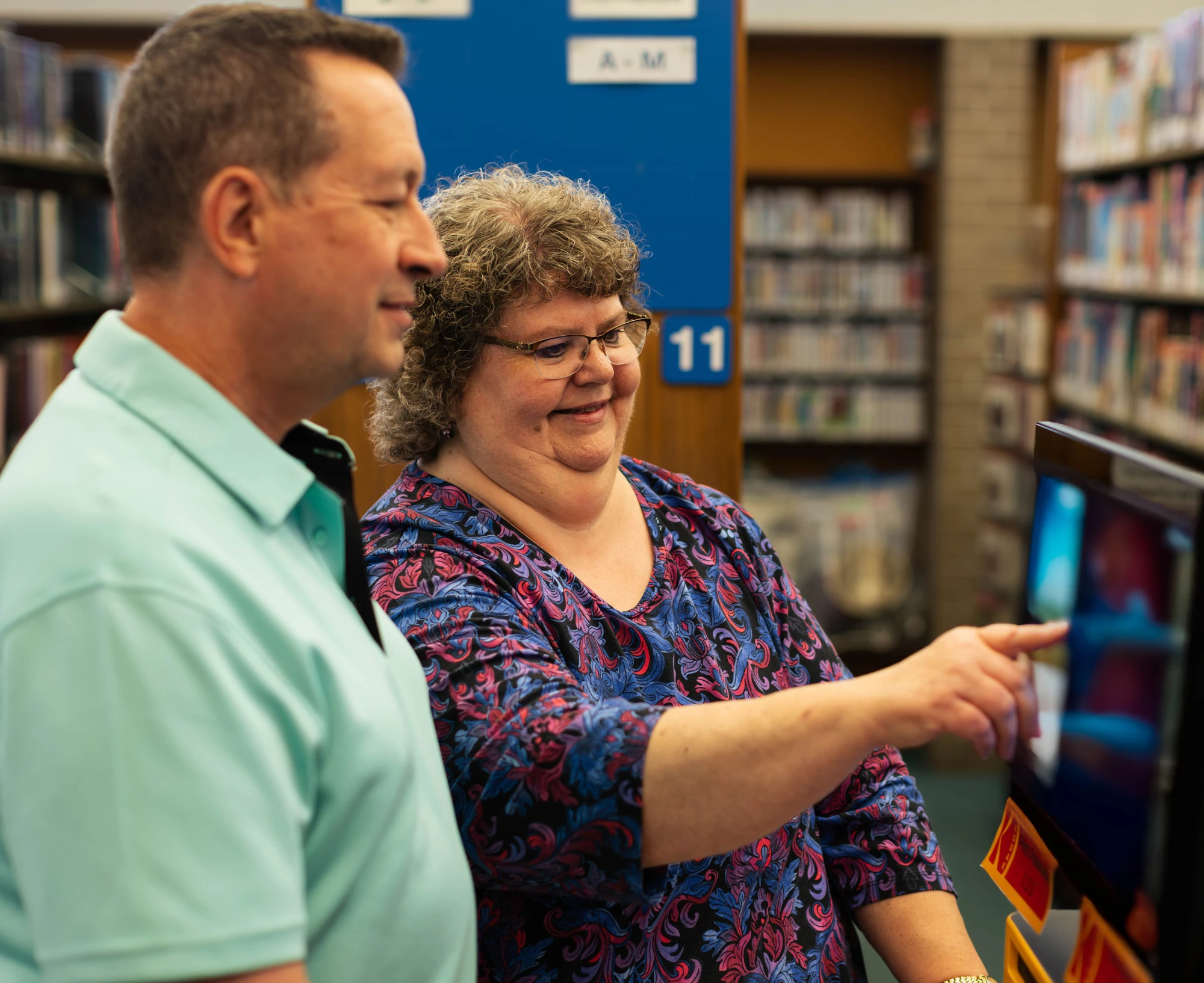 Two adults stand in a library with bookshelves in the background while one points to a screen.